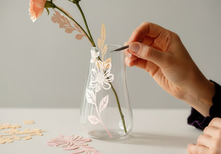 Close-up of a woman's hands carefully applying a delicate floral paper cutout to a curved glass vase.の写真素材