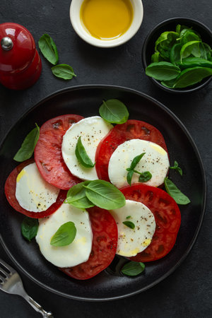 Caprese salad with fresh tomatoes, mozarella cheese, basil and olive oil on bkack plate, top view. Dark backgroundの写真素材