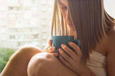 Sad young woman drinking tea, sitting on the window