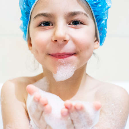 Little girl cheerfully takes a bath in the shower cap and plays with foamの写真素材