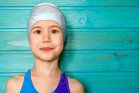 A seven-year-old girl in a blue swimsuit and a swimming cap against a blue wooden wall
