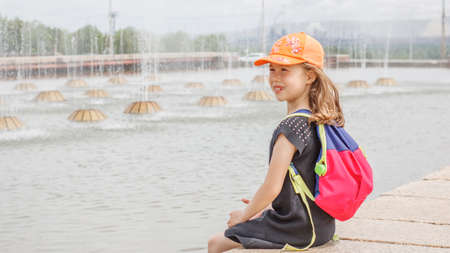 A girl of primary school age with a backpack behind her back walks by the city fountain. Summer...