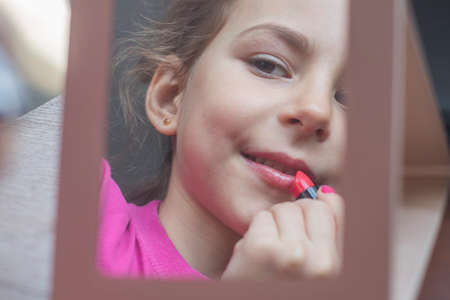 The little girl looks in the mirror and paints her lips with mom's lipstick.