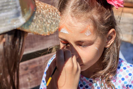 A girl paints a fox on her sister's face with watercolors, depicting face painting