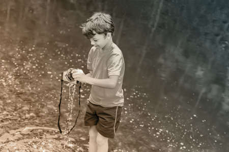 A boy in t-shirt and shorts is standing near the water in nature and washing a camera with soap and foam. The concept kid is a bully. Sepia, black and white photo.の写真素材