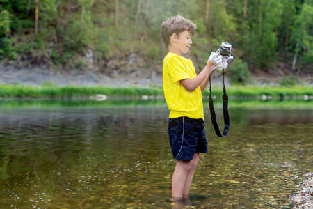 A schoolboy is standing in outdoor near the river, holding a photo camera in his hands and washing, cleaning it with soap and foam. The concept boy is a vandal. The risk of damage to equipment.の写真素材