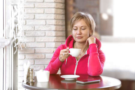A young thoughtful business woman in sportswear sits after a workout in a cafe and drinks coffee from a white Cup. The concept of lifestyle, success, beauty, healthy eating, and happiness.の写真素材