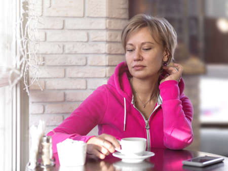 A young woman in a purple hoodie sits in a cafe and drinks coffee from a white cup. Daytime, lunch, portrait, close-up, soft light. Healthy lifestyle and sports concept. Indoors.の写真素材