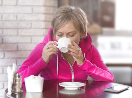 A young pretty woman in a purple hoodie sits in a cafe and drinks coffee from a white cup. Daytime, lunch, portrait, close-up, soft light. Healthy lifestyle and sports concept.の写真素材