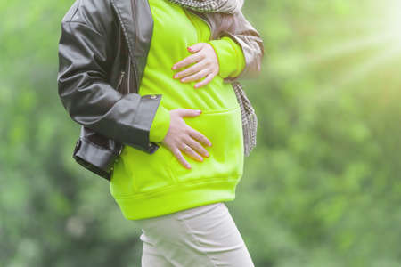 Pregnant woman dressed a green hoodie and black jacket holds hands on belly on a green background. Pregnancy, maternity, preparation and expectation concept. Close-up, outdoors. Nature, sunlight.の写真素材