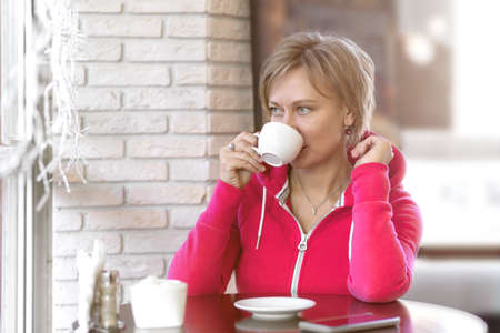 A young business woman in sportswear sits after a workout in a cafe, looks out the window at the street and drinks coffee from a white Cup. The concept of lifestyle, healthy eating, and happiness.の写真素材