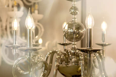 The picture of a glowing chandelier with metal chrome fixtures and shades in the form of candles. The concept of the classical style in the interior. Close up. Soft warm filter, selective focus.の写真素材