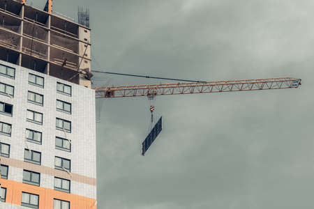 One yellow high-rise crane hauling plate against a house and gray sky during the construction phase. Industry concept for low-income young families. Mortgage, business, real estate loan. Copy space.の写真素材
