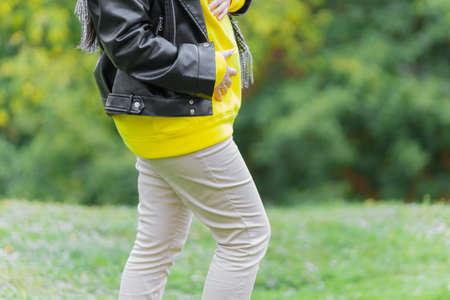 Pregnant woman dressed a hoodie and black jacket holds hands on belly on a green background. Pregnancy, maternity, preparation and expectation concept. Close-up, outdoors. Nature, sunlight.の写真素材