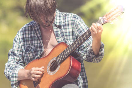 Adult male musician in a plaid shirt is playing the guitar on nature. Bright, sunny daytime, summer,...