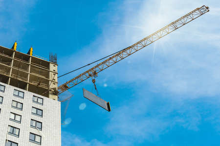 One yellow high-rise with glare crane hauling plate against a house and sunny blue sky during the construction phase. Copy space. Bottom up photo.の写真素材