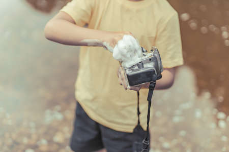 A boy in a yellow t-shirt and shorts is standing near the water on outdoor and cleaning a camera with soap and foam. The concept photographers day. Toned photo. Close up.の写真素材