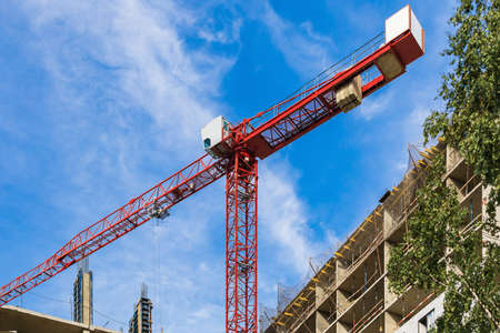 One high-rise crane against a house and sky during the construction phase. Industry concept for low-income young families. Mortgage, business, real estate loan. Summer. Green tree.の写真素材