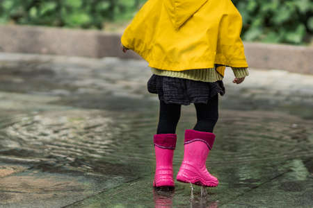 A nice little girl in a yellow raincoat and pink rubber boots jumps on puddles with splashes and rejoices. Park, nature, outdoors. Summer, warm. Universal Children's Day.の写真素材
