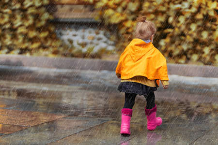 A happy little girl in a yellow raincoat and pink rubber boots walking in the rain on the park alone. Nature, outdoors. Childhood concept. Universal Children's Day.の写真素材