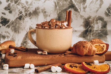 a cup of Christmas New Year delicious hot chocolate and cocoa with marshmallows sprinkled with cocoa powder on the wooden board, shape of spoon, dry orange slices, cones and croissants, cinnamon on a gray table, selective focus, hot drink winter or autumnの写真素材