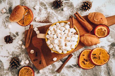 a cup of Christmas New Year delicious hot chocolate and cocoa with marshmallows sprinkled with cocoa powder on the wooden board, shape of spoon, dry orange slices, cones and croissants, cinnamon on a gray table, selective focus, hot drink winter or autumn top viewの写真素材