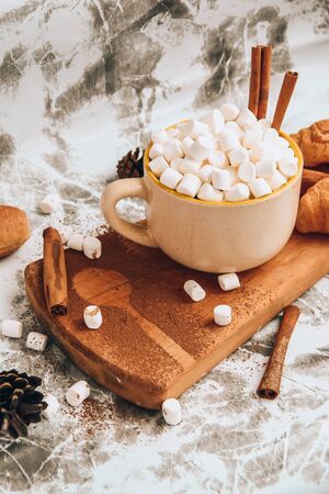 a cup of Christmas New Year delicious hot chocolate and cocoa with marshmallows sprinkled with cocoa powder on the wooden board, shape of spoon, cones and croissants, cinnamon on a gray table, selective focus, hot drink winter or autumnの写真素材