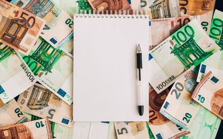 Opened spiral notepad sheet in a cage on money background, top view, copy space, A white notebook with blank pages lay on a set of banknotes of 100 euros, 50 euros, 20 euros 10 eurosの写真素材