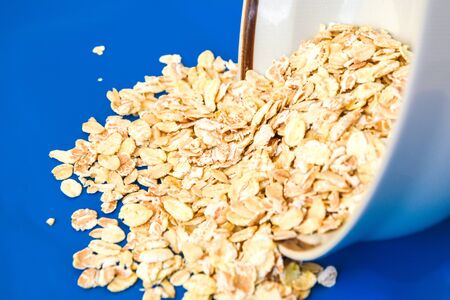 Uncooked oatmeal spilling from a ceramic bowl, on blue background, ingredient for healthy breakfast,...