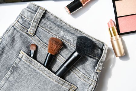 Different makeup products composition with jeans on white background, flat lay, top view, eyeshadow, lipstick, highlighter, make up brushes on jeans pocketの写真素材