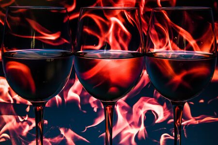 transparent glasses of wine on fire flame background, Three wine glasses flutes in a row with colorful multicolored light painting behind them, copy spaceの写真素材