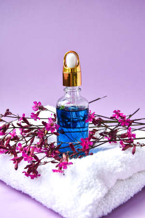 Glass dropper with a drop of cosmetic oil and dried flowers and herbs on purple background. Natural organic herbal skin care oil, white towel, small pink flowersの写真素材