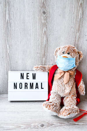 Coronavirus covid-19. Teddy bear doll wearing mask backpack and holding lightbox with text NEW NORMAL on wooden background, copy space, Quarantine 2020. Back to schoolの写真素材