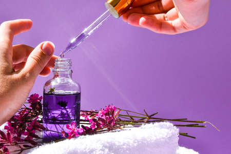 Woman dripping rose essential oil on hand against blurred flowers on pink background, closeup, white spa towel, beauty treatment, hand skin care, natural medicine, Women's hands applying oil with a dripの写真素材