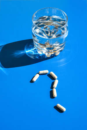Glass of water and pills in shape of question mark on blue background. Hard light and shadows. Vitamins and prebiotics, probiotics. Autumn vitamin dose.の写真素材