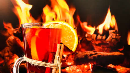 Hot mulled wine in a glass next to the burning fireplace. Hot christmas beverage. Orange slices, cinnamon sticks, anise star. Copy space. Holiday atmosphere. Christmas greeting cardの写真素材