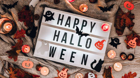 HAPPY HALLOWEEN. Text in white light box, with a background of sweater, dried leaves halloween autumn decoration. Ghost, pumpkins bat, black cat. Home decoration. Top view. Bokeh lightの写真素材