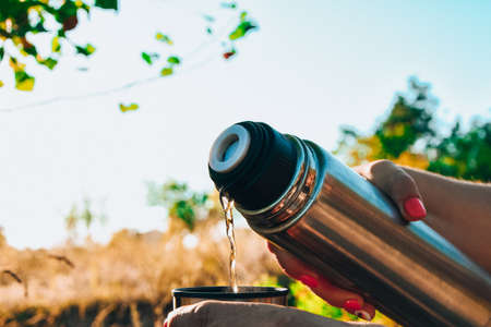 Traveler pouring hot tea from flask to cup at autumn fallen forest outdoor. Summer autumn picnic. Copy spaceの写真素材