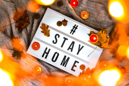 Hashtag Stay home. Text in white light box, with a background of sweater, candles, dried leaves autumn decoration. Coronavirus devices. Social distancing. Protection against pandemics. Stay safe. Cozy homeの写真素材