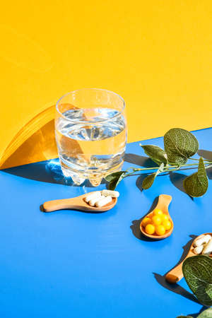 Glass of water and pills on wooden spoon on blue and yellow background. Hard light and shadows....