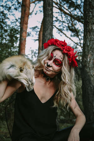 Happy Halloween concept. Scared horrible female sitting in special outfit, wears white clay skull, professional sugar skull makeup to look spooky, wears black clothes playing with fox fur. Day of The Dead. Dia de los muertos.の写真素材