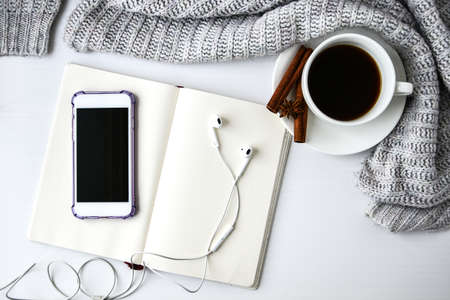 Cup of coffee with notebook phone headphones cinnamon sticks and anise star on white background. Sweater around. Winter morning routine. Coffee break. Copy space. Top view. Breakfast. Flat layの写真素材
