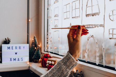 Female hand draws christmas holidays decoration painted on window glass. New Years scenery. city buildings garlands lights. hygge, decoration and christmas concept. festive garland string at homeの写真素材