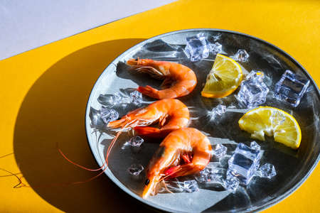 Red shrimps with lemon and ice on a gray plate and yellow background. Trendy colors. Minimalistic concept in isometric style. Sea food. Vegetable omegaの写真素材