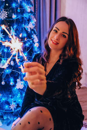 Happy young woman sitting with glass of champagne and bengal light next to the christmas tree. New...