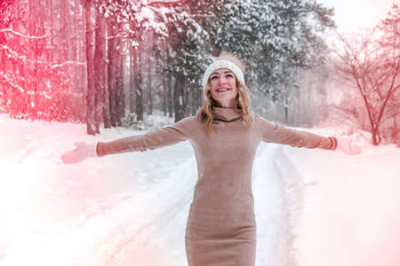 Christmas, holidays and season concept. Young happy woman blowing snow in the winter forest nature. Warm clothing knitted gloves and hat. Winter forest landscape backgroundの写真素材