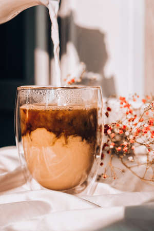Milk pouring into the glass cup with black coffee. White cup of milk and chocolate. Morning aesthetics vibes. Breakfast. Pink gypsophila flowers. Hard shadows.の写真素材