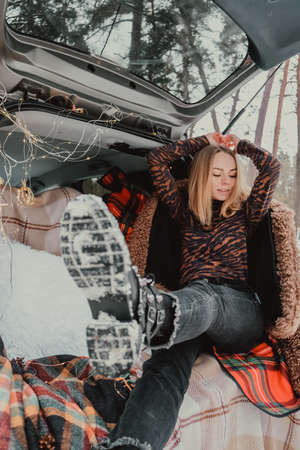 Blonde woman wrapped in blanket in trunk car. Travel in winter. Car decorated with festive Christmas...
