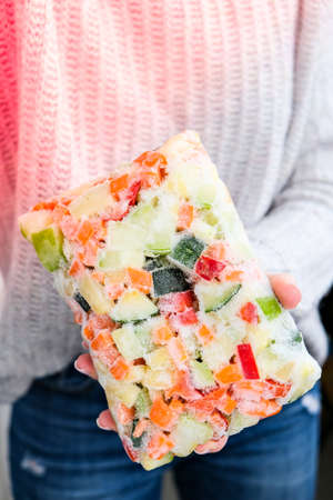 Mixed vegetable frozen food briquette in female hands. Preparing harvest for winter. Veganism, vegetarian healthy eating. Ice with foodの写真素材