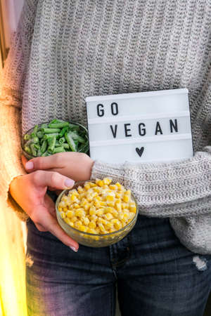 Lightbox with text GO VEGAN in female hands. Veganism, vegetarian healthy lifestyle. Frozen food in bowl. Healthy eating vegan. Green beans and yellow cornの写真素材